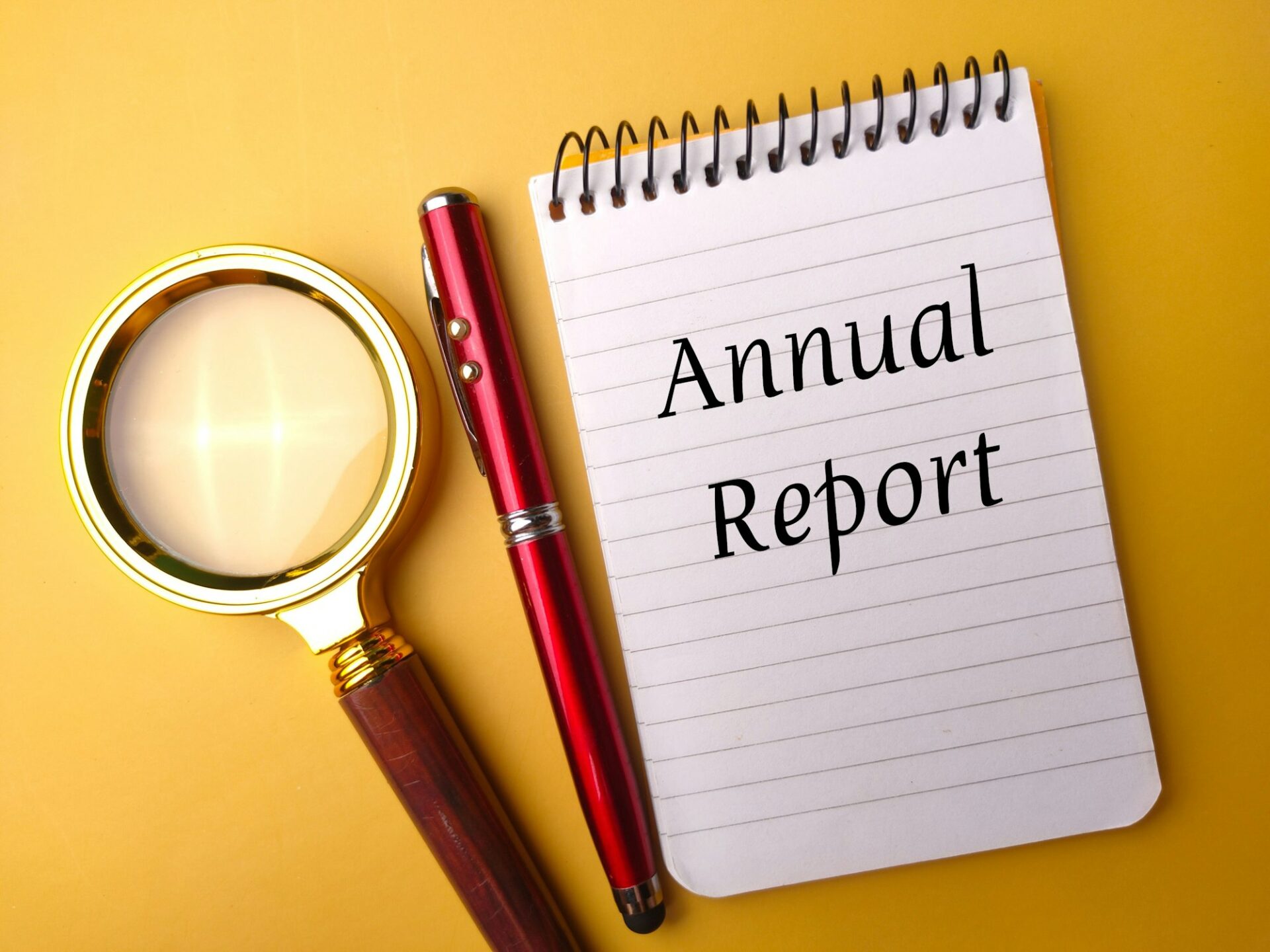 Closeup of a notepad with the text "annual report" next to a pen and a magnifying glass