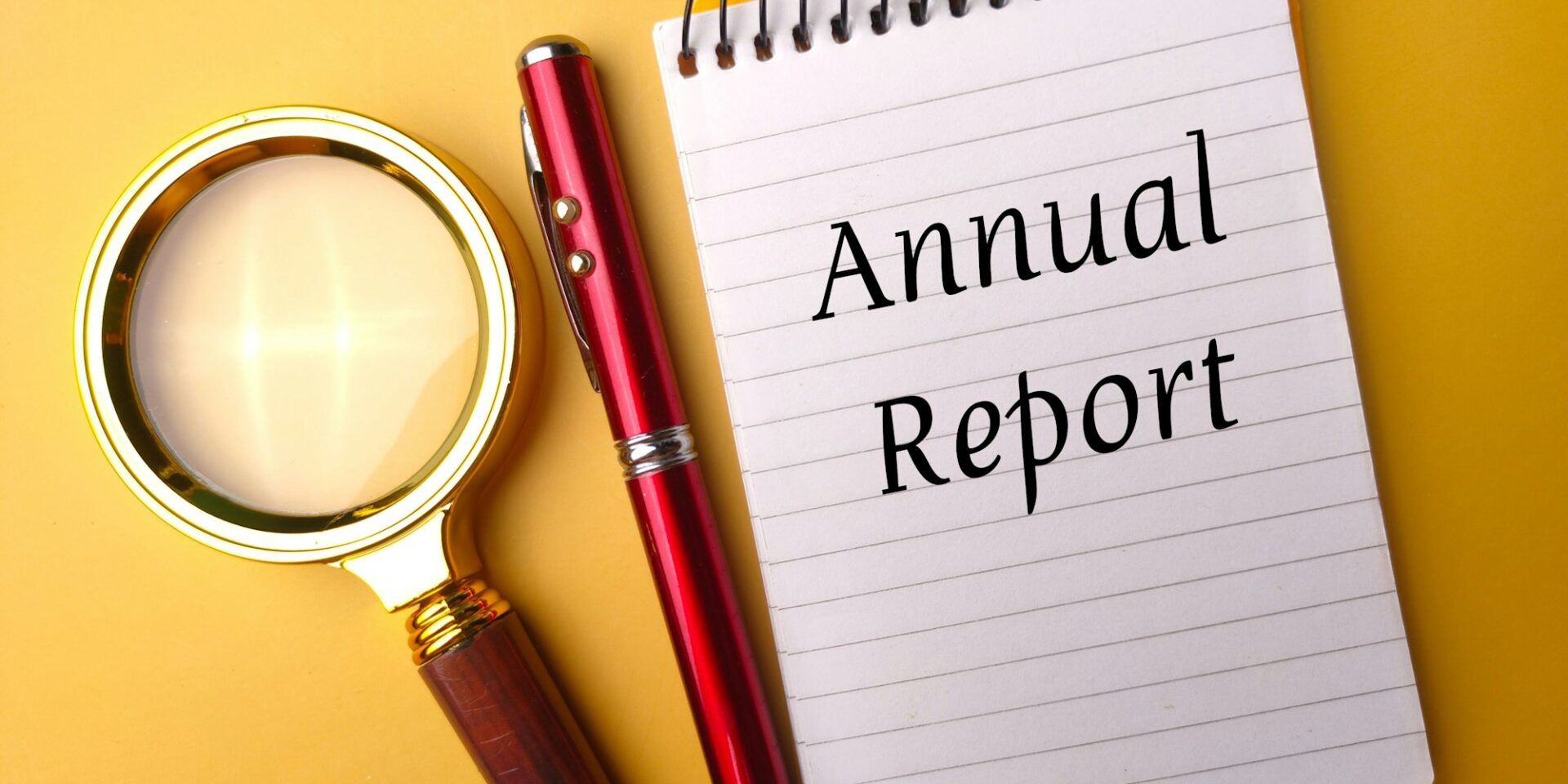 Closeup of a notepad with the text "annual report" next to a pen and a magnifying glass